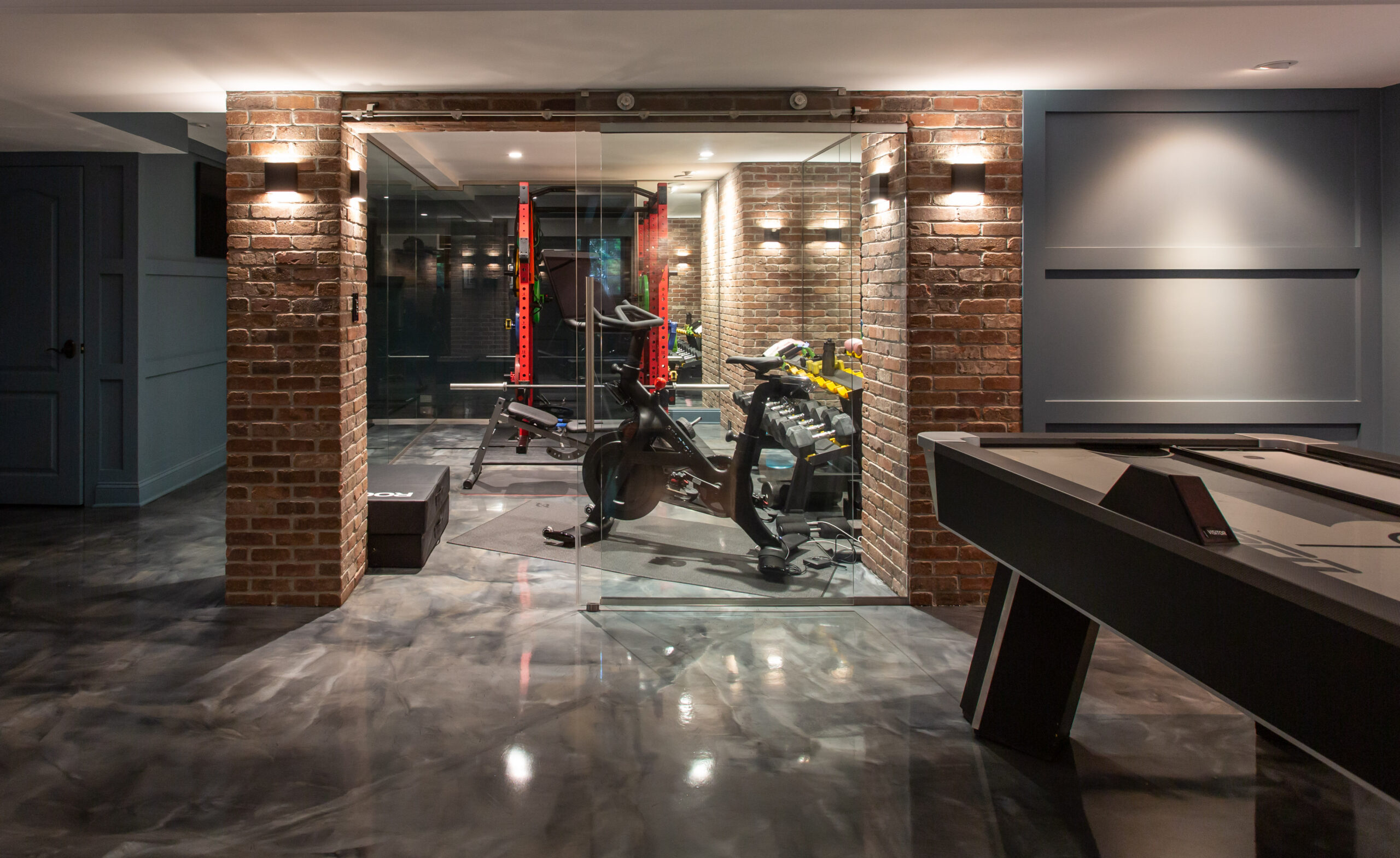 Modern home gym with brick walls and glass enclosure, adjacent to a game table.