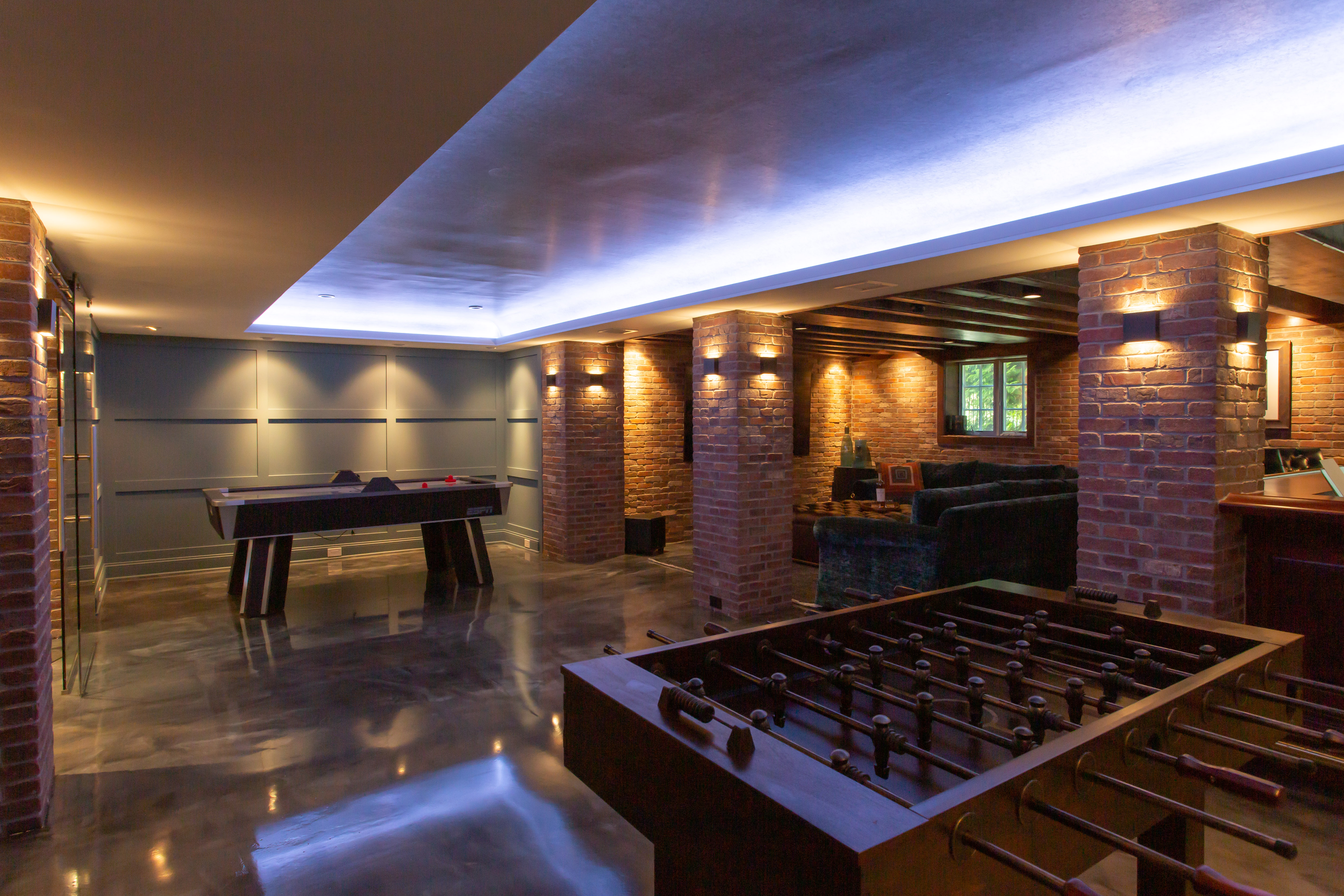 Modern game room with foosball and air hockey tables, illuminated by warm brick wall lights.